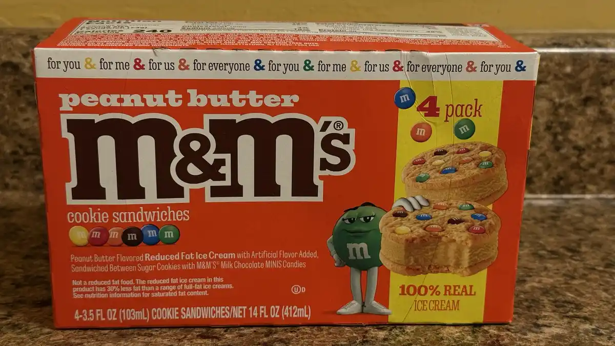 A box of peanut butter M&M's cookie sandwiches sits on a countertop. The box features a green M&M character and images of…