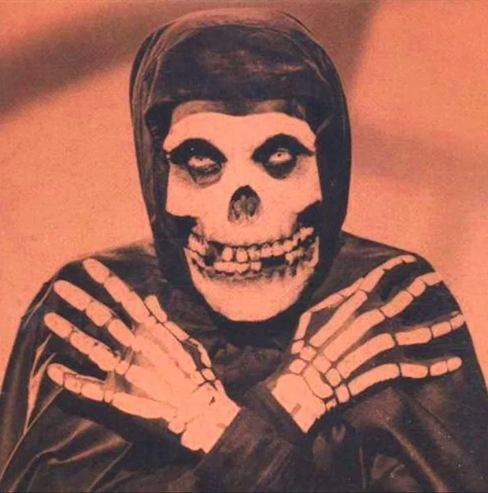 A person dressed as the grim reaper with a skull face, hooded robe, and skeletal hands clasped together in front of their…