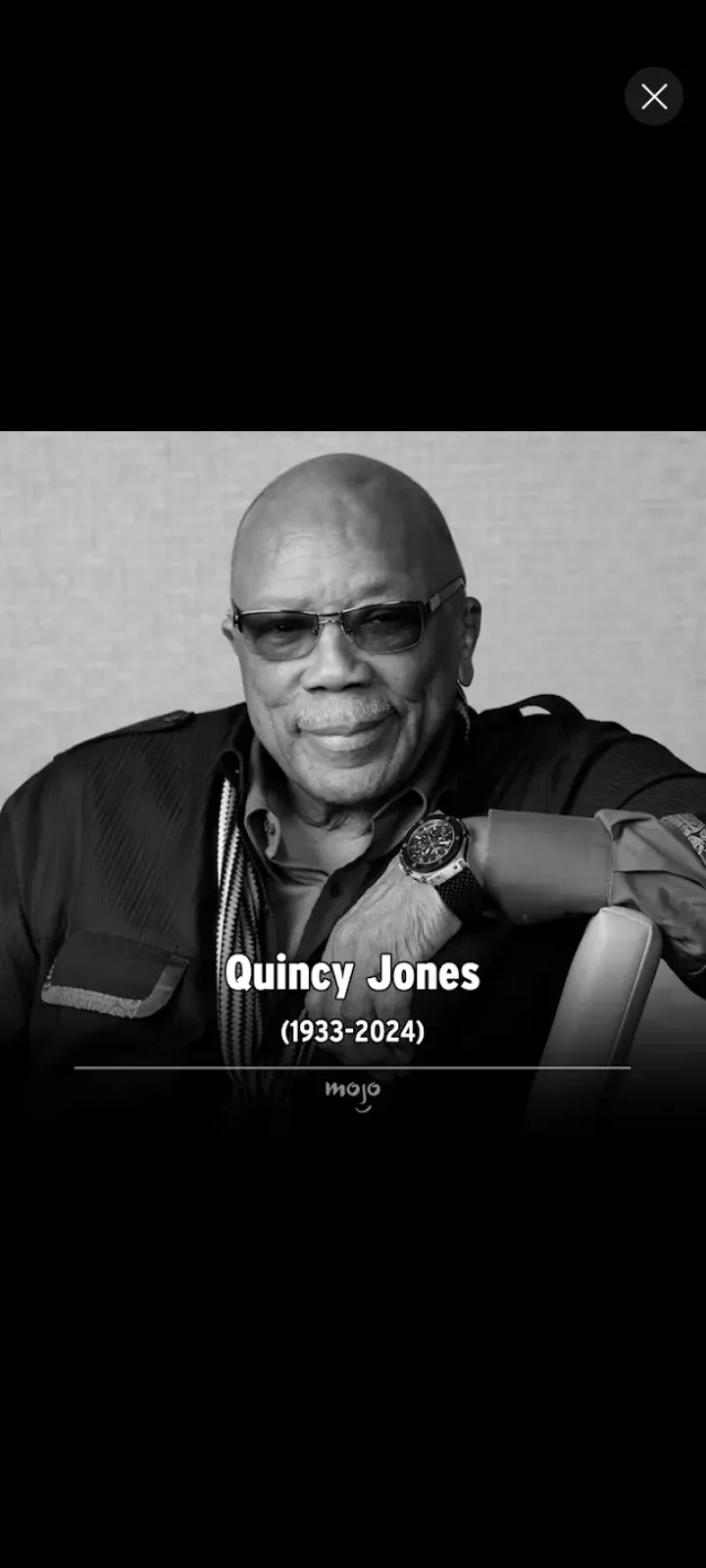 A black and white portrait of Quincy Jones, a bald man wearing glasses and a dark jacket, smiling while resting his arm on a…