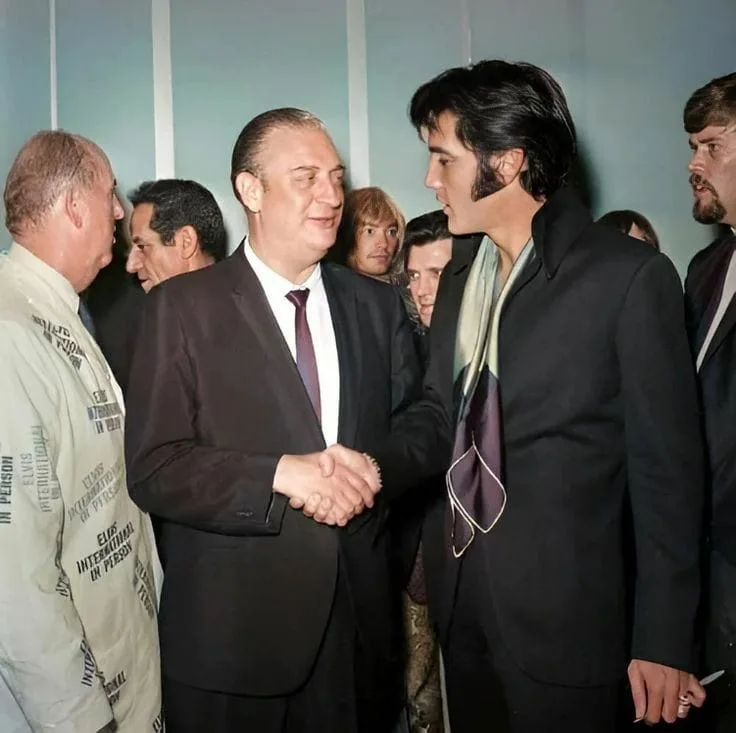 A historical photograph shows Elvis Presley shaking hands with a man in a suit, surrounded by other people. Elvis is wearing…