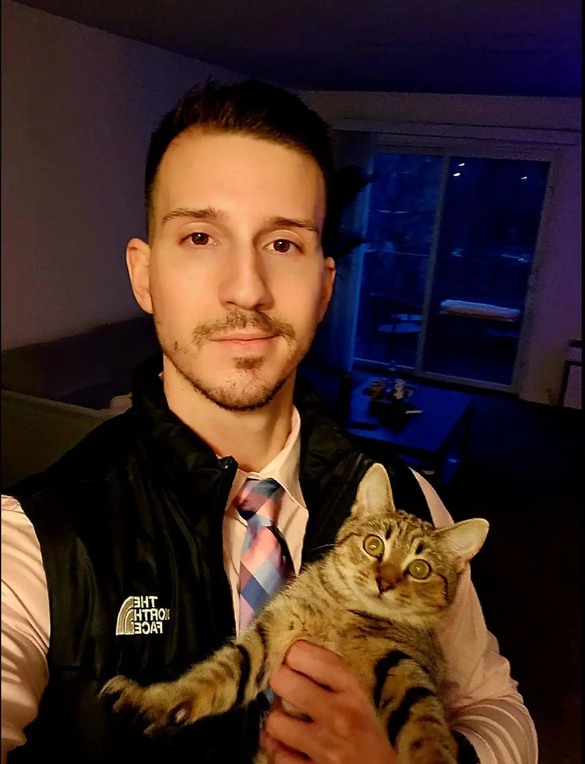 A man in a black vest over a collared shirt and tie holds a tabby cat. The cat has wide, startled eyes and looks directly at…