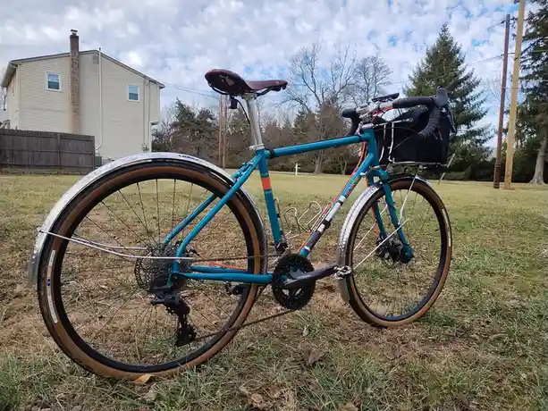 A teal bicycle with brown tires and silver fenders is parked on grass in front of a house, featuring a black bag on the…