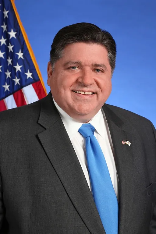 A smiling man in a dark suit and blue tie stands before a blue backdrop with an American flag to his left. He wears a small…