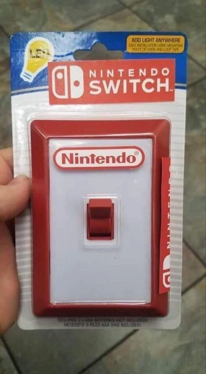 A hand holds a light switch in its retail packaging, which is designed to look like a Nintendo Switch box. The packaging…