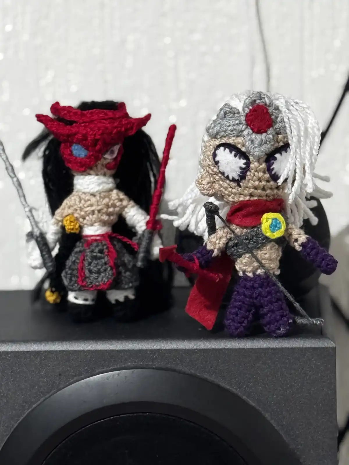 Two crocheted figurines of video game characters stand side by side on a speaker. The character on the left has a red mask…