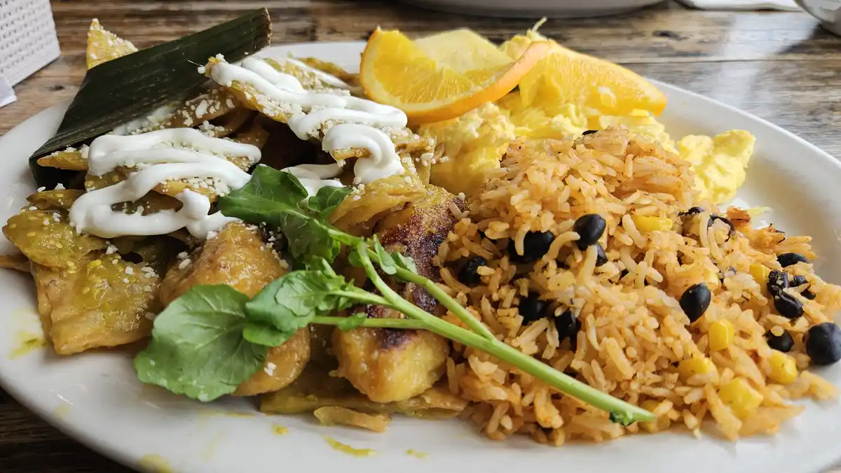 A plate of food featuring fried rice with black beans and corn, scrambled eggs, orange slices, and what appears to be…