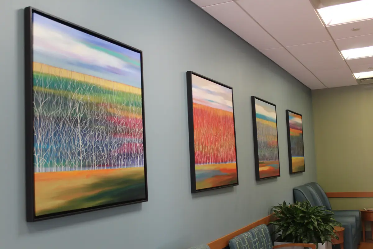 A row of four colorful abstract paintings depicting stylized trees and landscapes hangs on a gray wall in a waiting room…