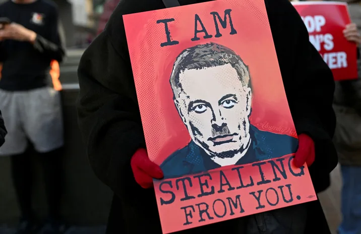A person wearing a black jacket and red gloves holds a red protest sign featuring a stylized, halftone portrait of Elon…