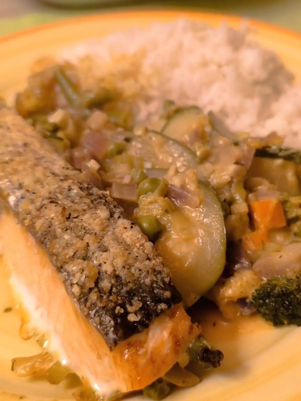 A close-up photo of a plate of food featuring a piece of fish with a textured, dark crust, served with mixed vegetables like…