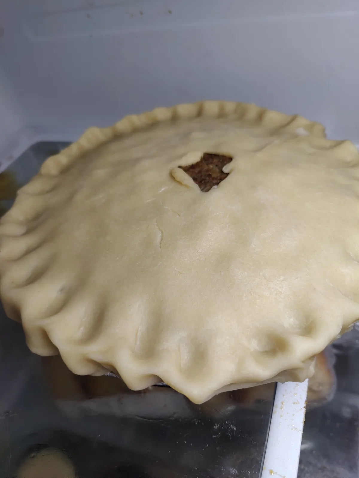 A close-up photo of an unbaked pie with a fluted crust and a small, irregularly shaped hole cut in the center, revealing the…