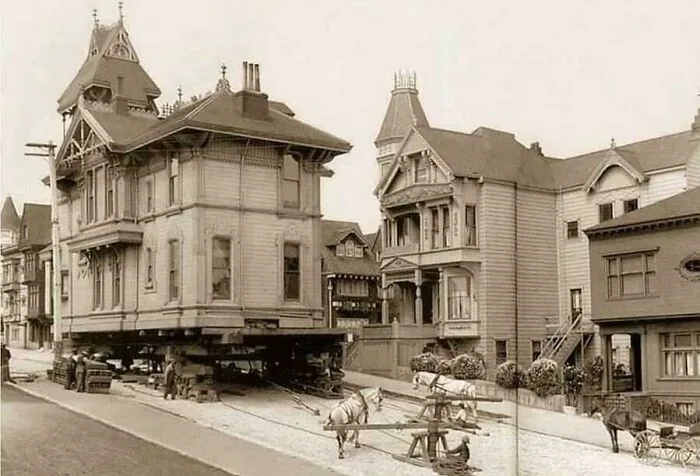 a sepia-toned historical photograph shows a large, ornate Victorian-style house being moved on a custom-built wooden dolly…