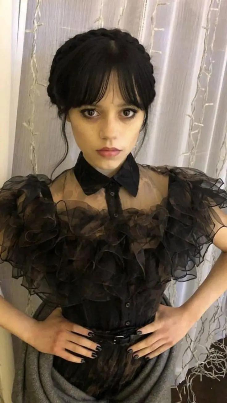 A young woman with dark hair styled in a braid and bangs, wearing dramatic black makeup and a sheer black ruffled blouse…