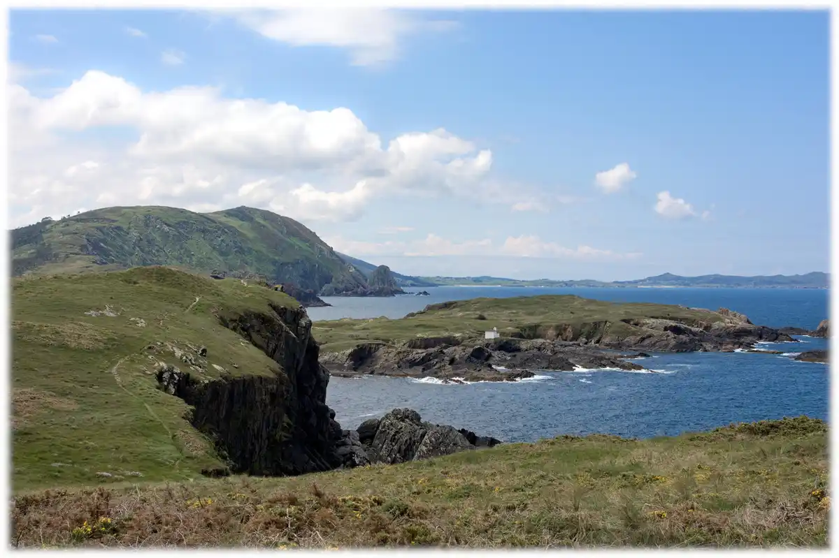 A scenic coastal landscape with green hills, rocky cliffs, and blue ocean under a partly cloudy sky. A small white building…