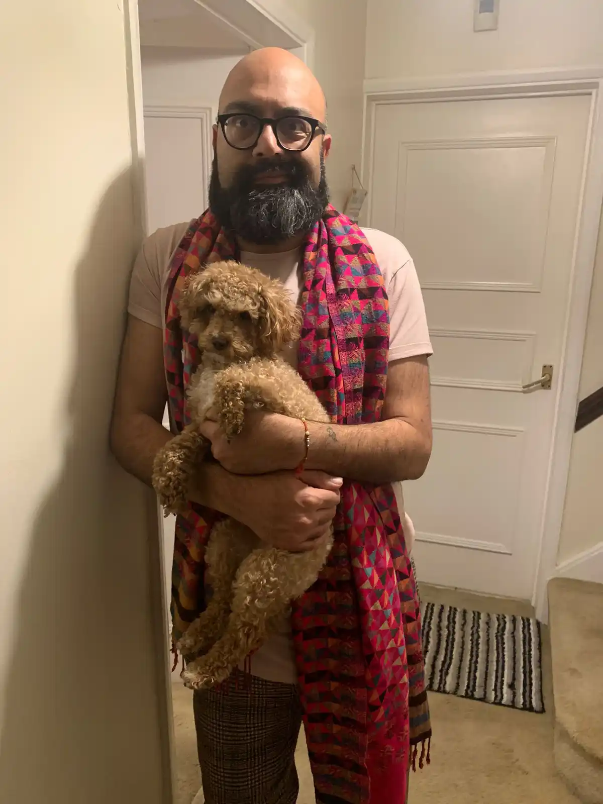 A bald man with a full beard and glasses, wearing a colorful geometric scarf and plaid pants, stands indoors holding a…