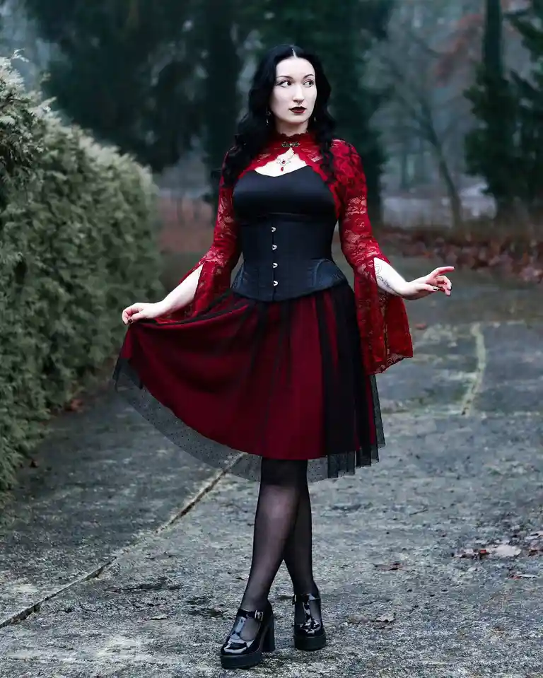 A woman in a gothic outfit with a black corset, red lace cape, and polka-dot skirt stands on a path, looking sideways with a…