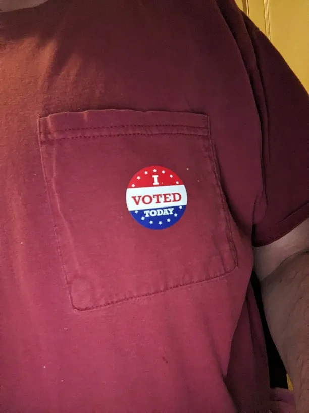 A close-up photo shows a red, white, and blue circular 'I VOTED TODAY' sticker pinned to the chest pocket of a maroon…