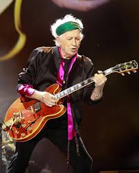 A man with white hair and a green headband plays a red electric guitar on stage. He wears a black jacket over a purple…