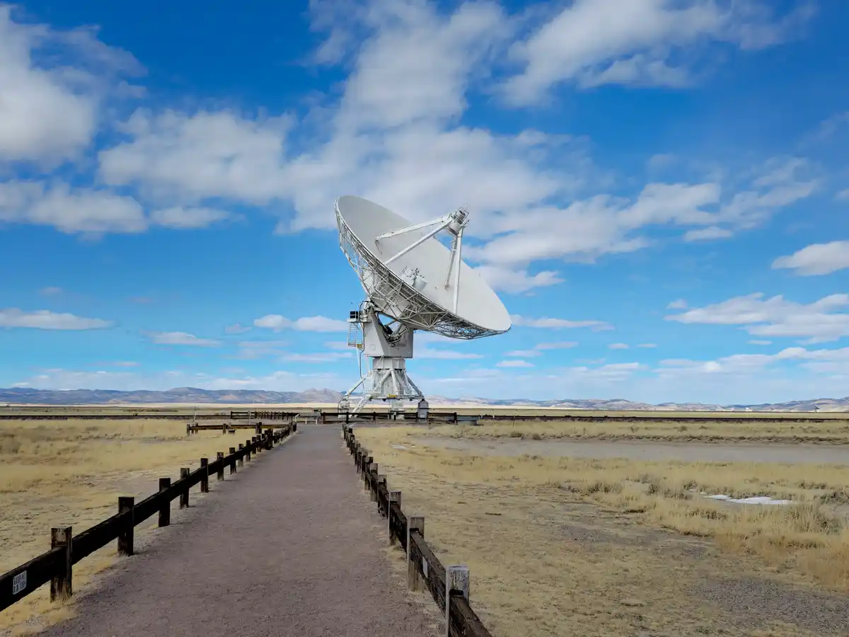 A large white radio telescope stands in a vast, arid desert landscape under a bright blue sky with scattered clouds. A…