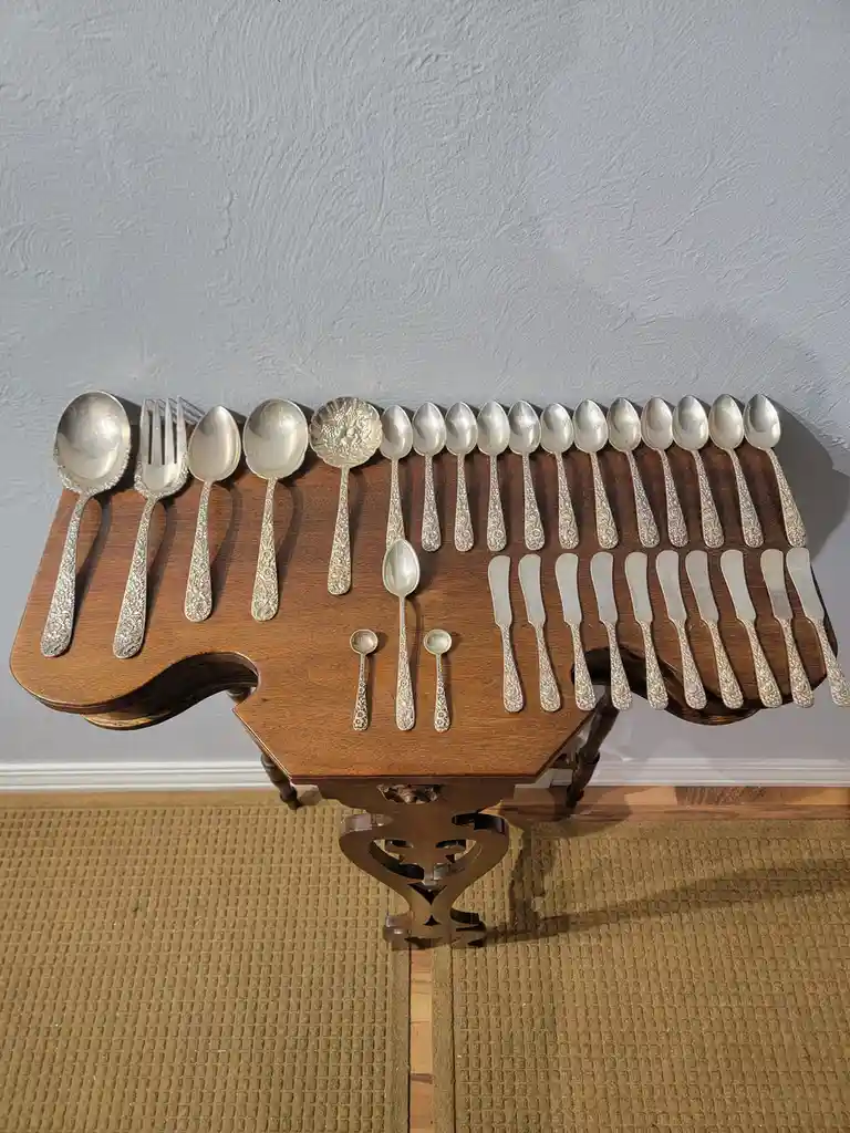 A wooden serving tray holds an extensive collection of ornate silverware, including multiple spoons, forks, and butter…