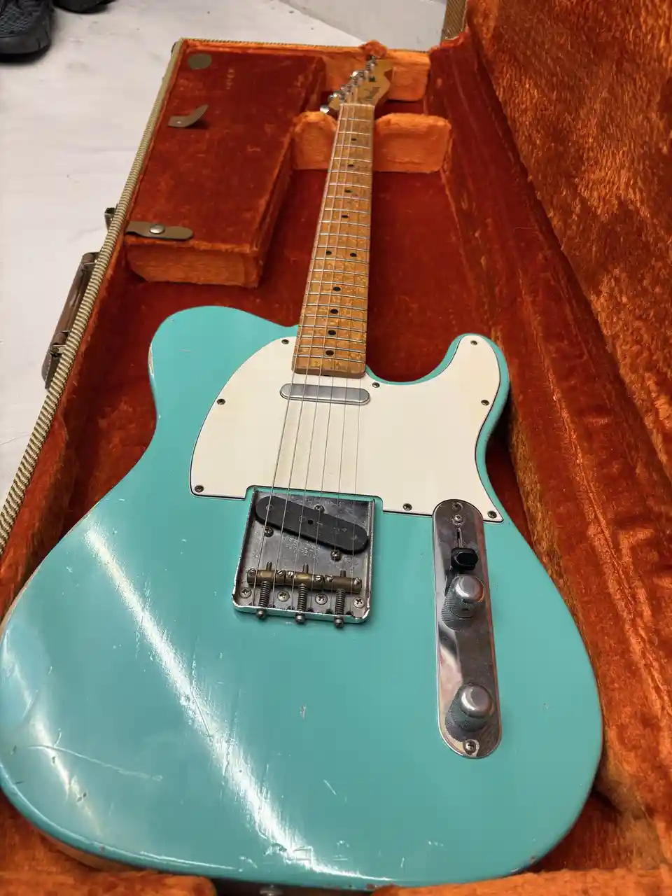 A turquoise electric guitar with a white pickguard and worn finish sits inside an orange velvet case. The guitar's bridge is…