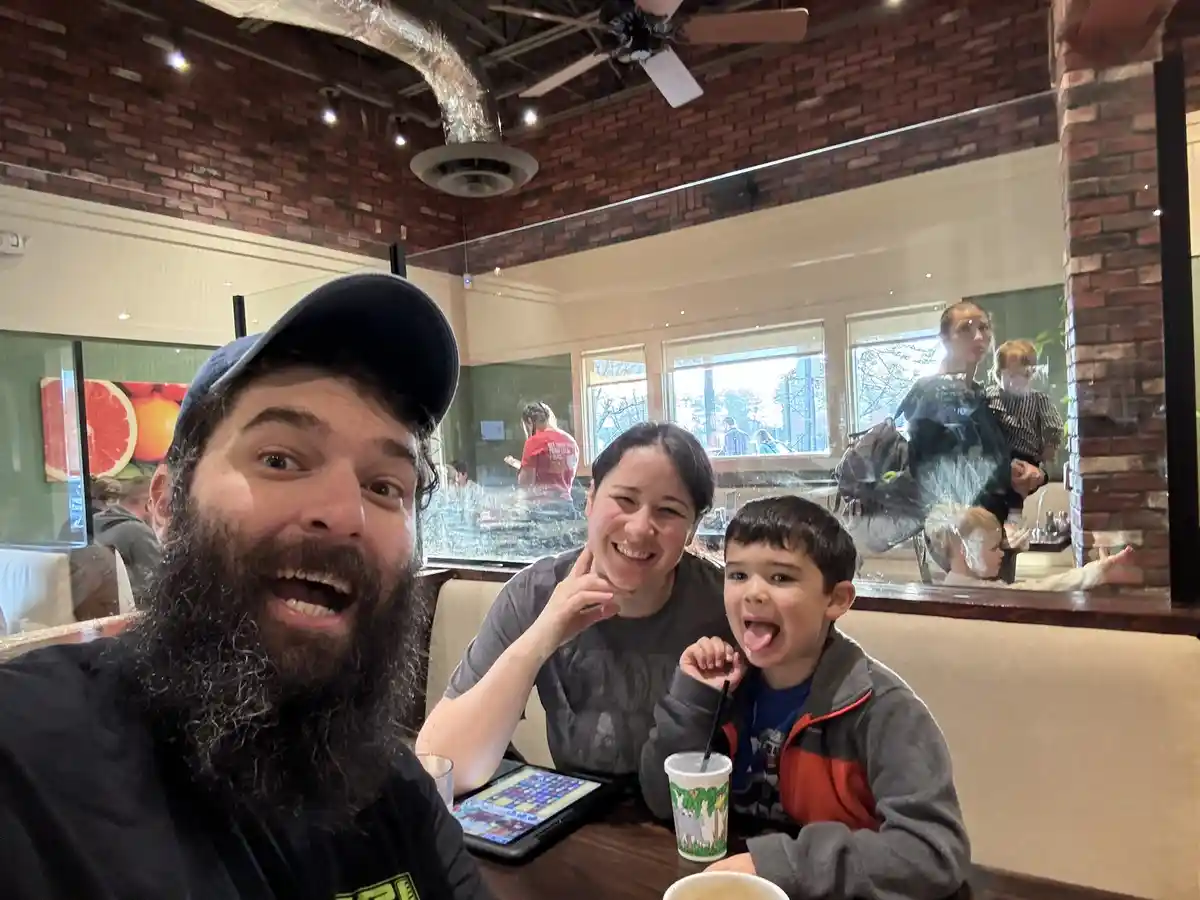 A family photo shows a bearded man in a cap, a smiling woman, and a young boy sticking his tongue out, all seated at a…