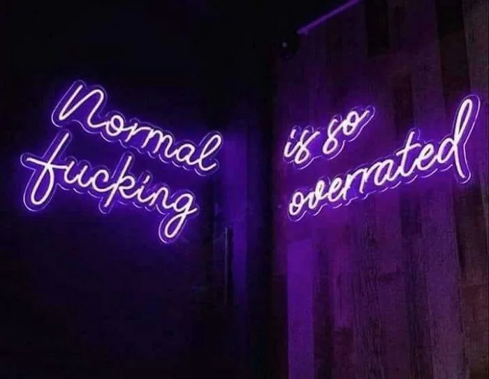 Two purple neon signs on a dark wooden wall read 'Normal fucking' on the left and 'is so overrated' on the right, conveying…