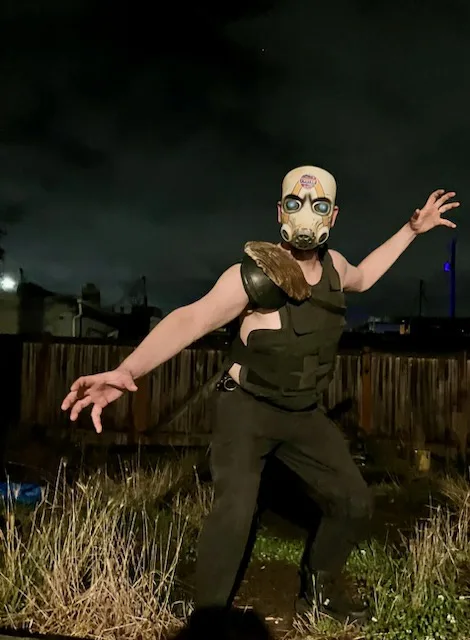 A person wearing a gas mask and tactical vest stands outdoors at night with arms outstretched, holding a chicken wing. The…