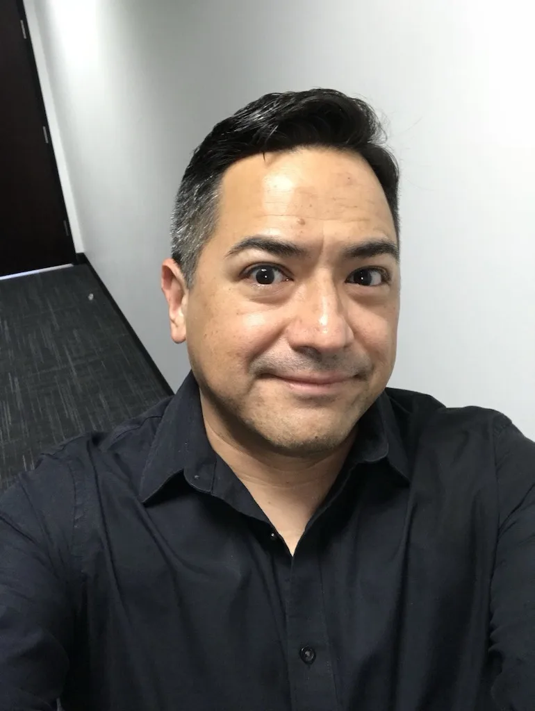 A man with dark hair and a black button-up shirt looks directly at the camera with wide eyes and a subtle, knowing smirk…