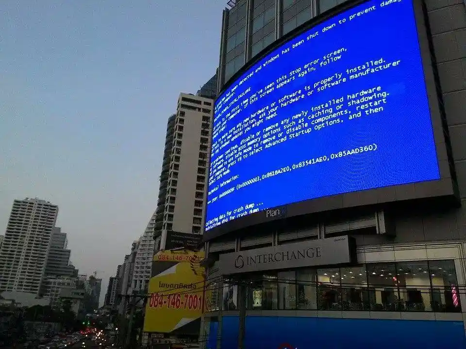 A large outdoor billboard in a city displays a blue Windows Blue Screen of Death (BSOD) error message, with text advising…