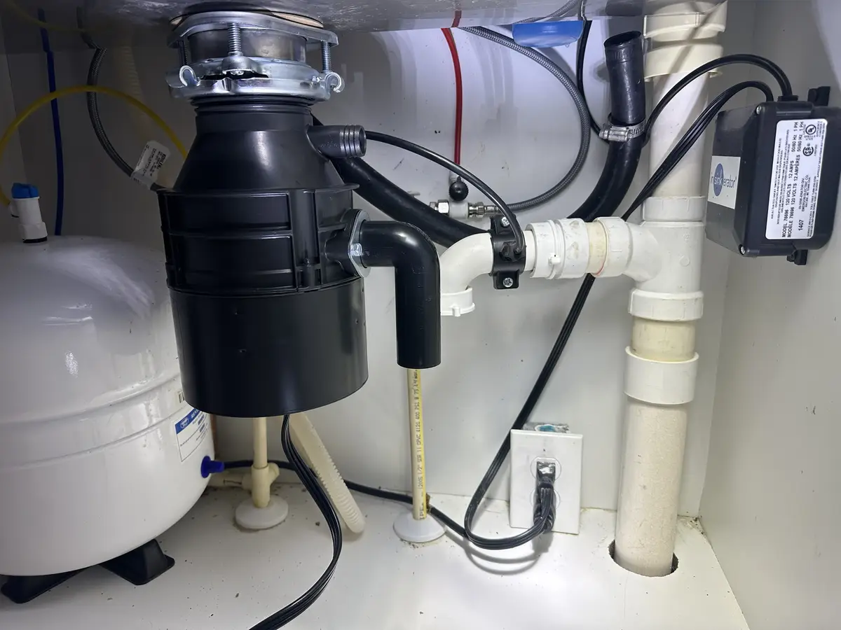 A close-up photo of a kitchen sink's under-counter plumbing, prominently featuring a black garbage disposal unit connected…