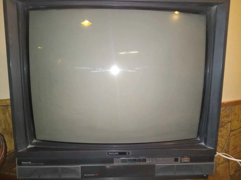 An old, bulky Philips CRT television with a blank, reflective screen showing light glare, sitting on a wooden surface with a…