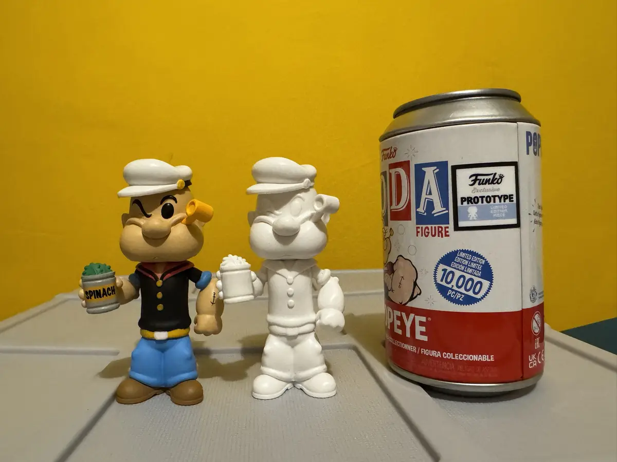 Two Funko Pop! figures of Popeye are shown side by side against a yellow wall. The left figure is fully colored, holding a…