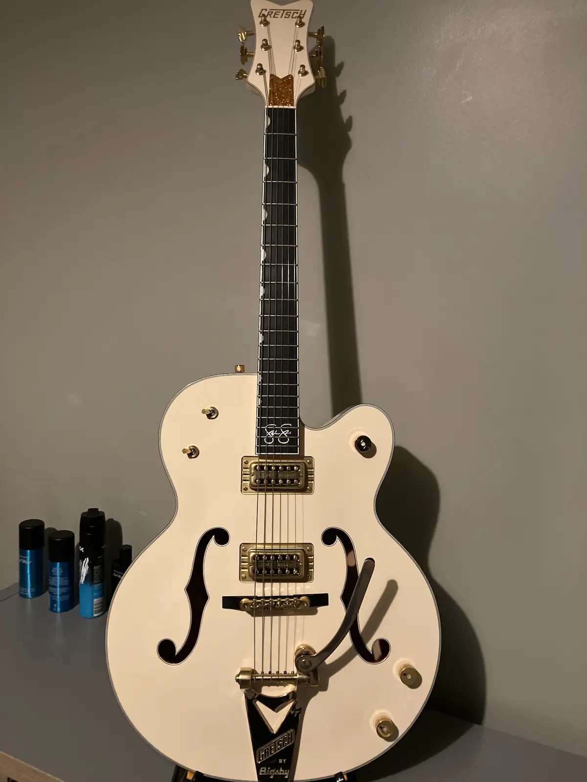 A white Gretsch electric guitar with f-holes and gold hardware stands upright against a plain wall, with several blue and…