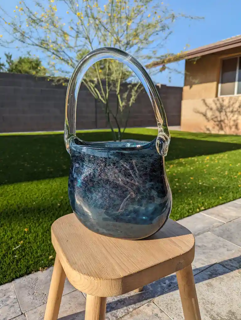 A handmade glass purse with a glittery blue interior and clear handle sits on a wooden stool outdoors on a sunny day, with a…