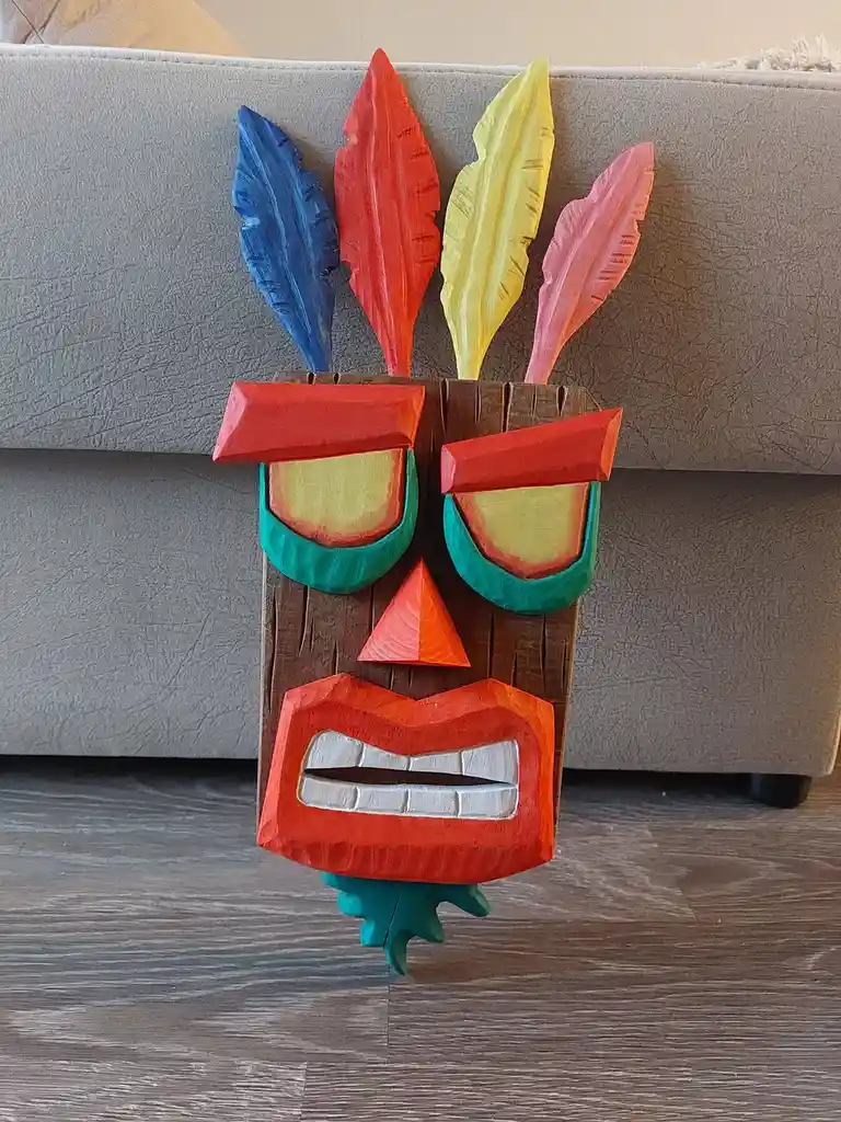 A colorful, hand-carved wooden tiki mask with four painted feathers (blue, red, yellow, pink) on top, standing upright on a…