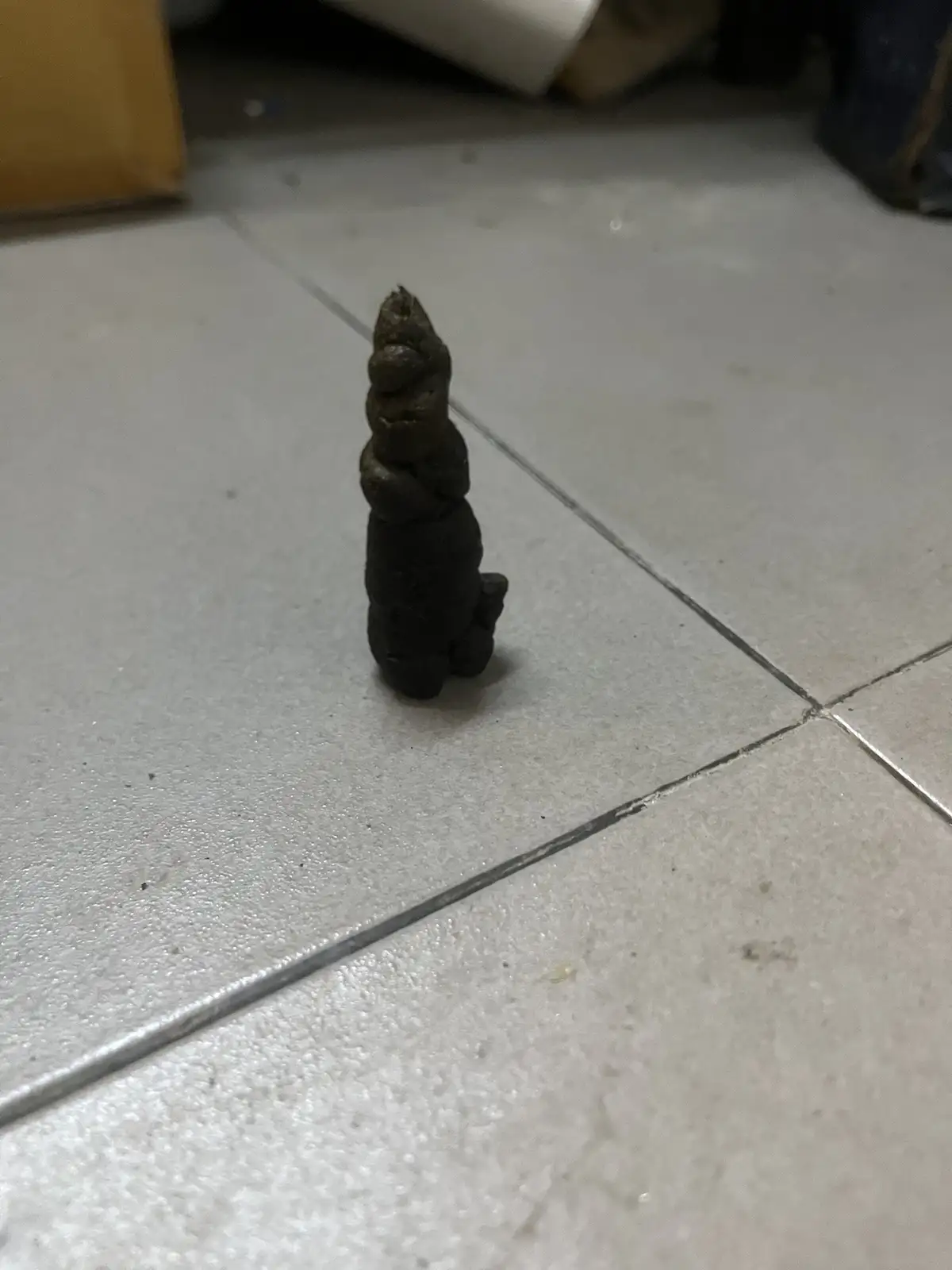 A dark, twisted, spiral-shaped piece of animal feces stands upright on a light-colored tiled floor, resembling a unicorn…