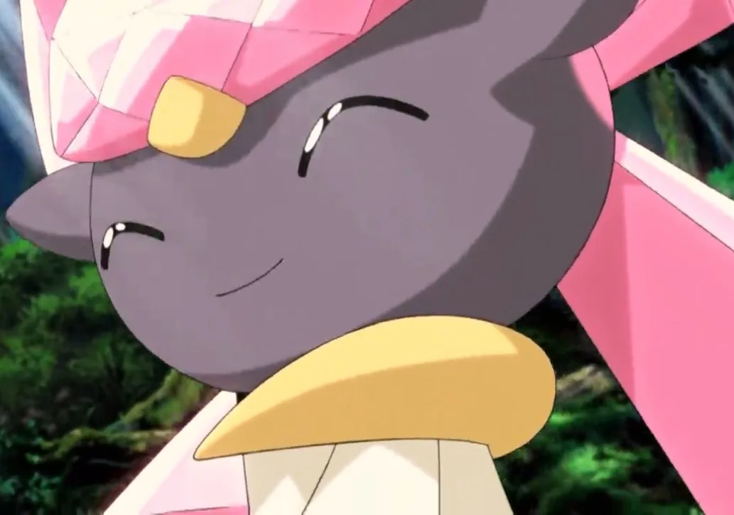 A close-up of the Pokémon Diancie, a grey cat-like creature with a pink diamond-shaped headpiece and yellow accents, smiling…