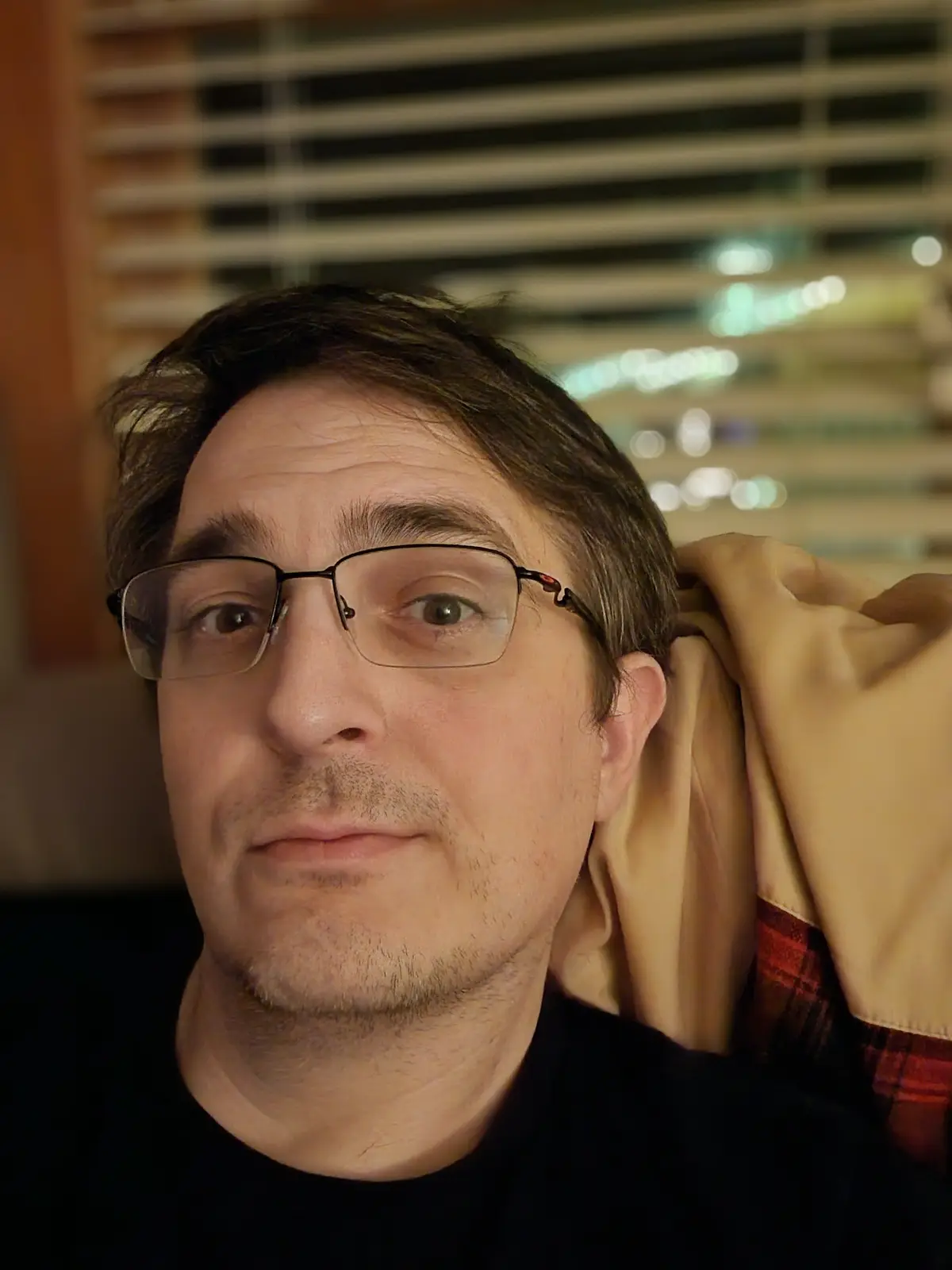 A man with glasses and a black shirt looks directly at the camera with a neutral expression while holding a beige jacket…