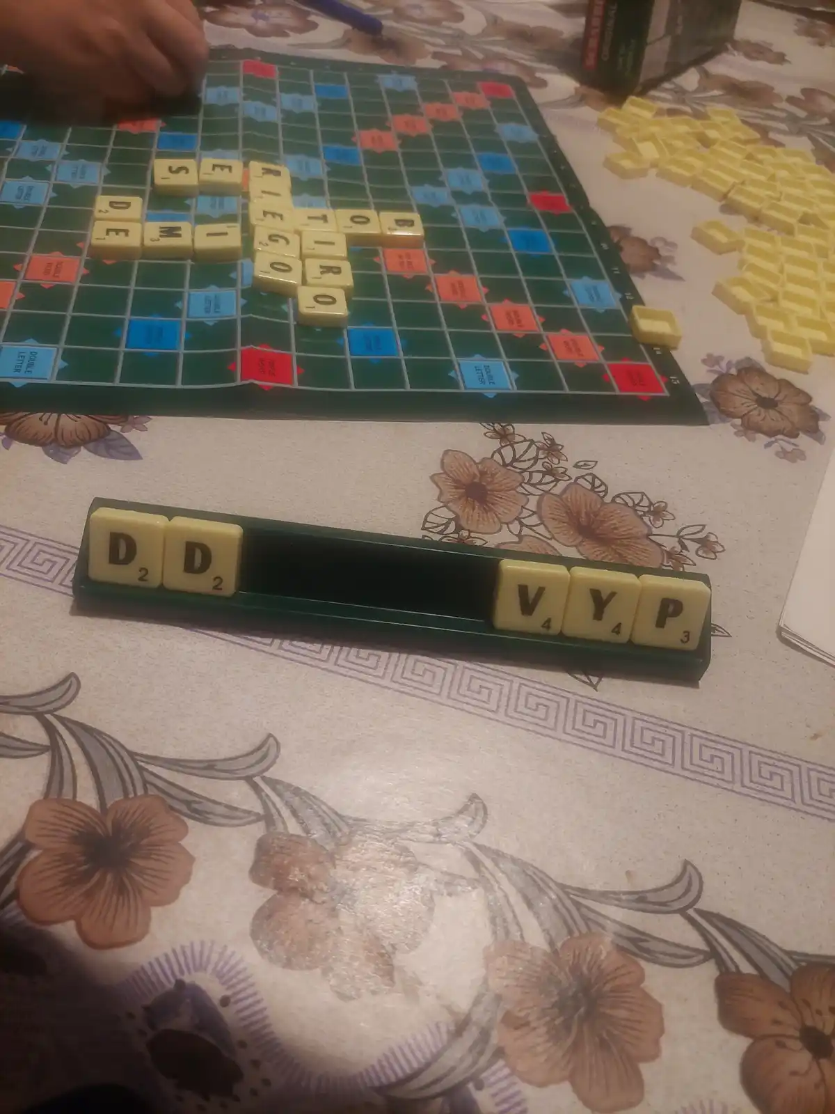 A close-up photo of a Scrabble game in progress on a floral-patterned tablecloth, showing a rack with the letters 'V', 'Y'…
