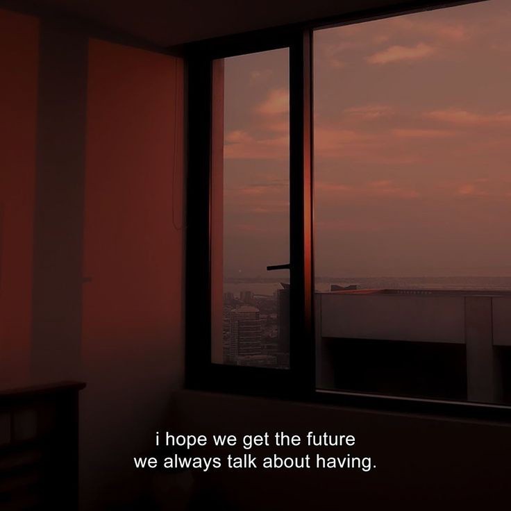 A dimly lit room with a large window showing a cityscape at sunset or sunrise, bathed in warm orange and pink hues. White…