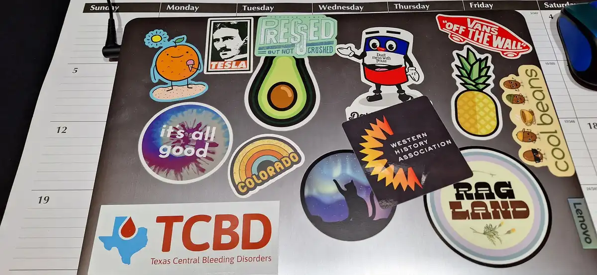 A close-up photo of a cluttered computer mousepad covered in various colorful stickers, including a cartoon orange, a Tesla…