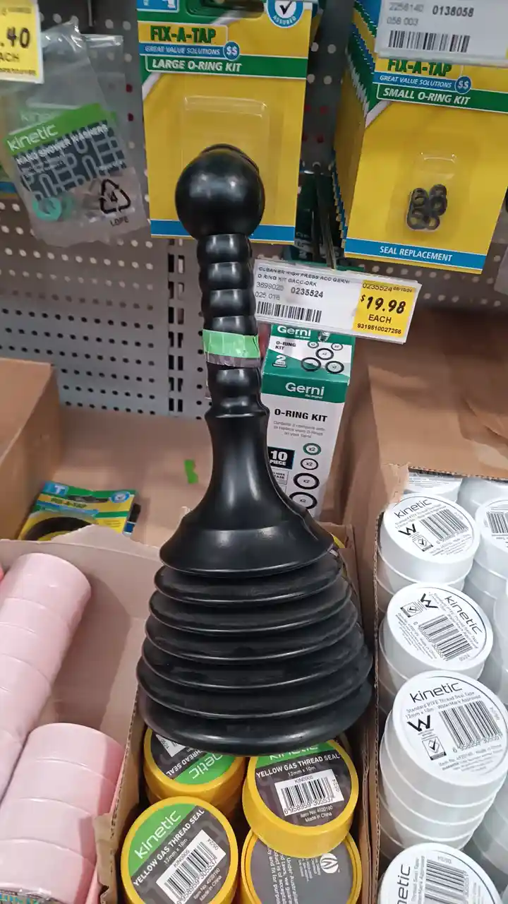 A large black plunger is prominently displayed in a retail box, surrounded by various plumbing supplies like 'Fix-A-Tap'…