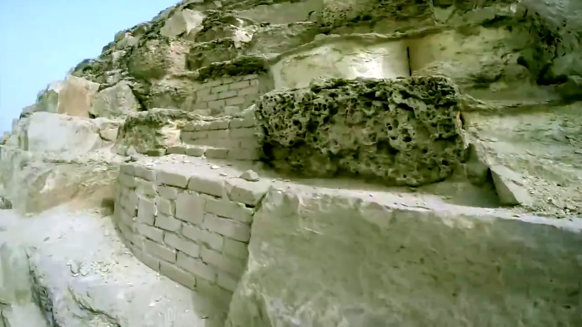 A close-up shot of ancient, weathered stone ruins with layered rock formations and a large, porous volcanic rock block…