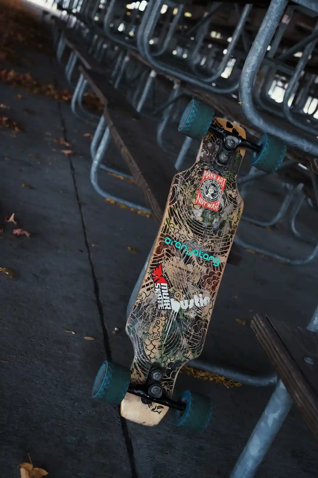 A longboard skateboard with a worn, graffiti-covered deck and teal wheels leans against a metal barricade in an outdoor…