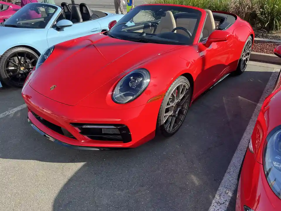 A shiny red Porsche 911 convertible is parked in a lot, with a light blue Porsche 911 convertible behind it and another red…