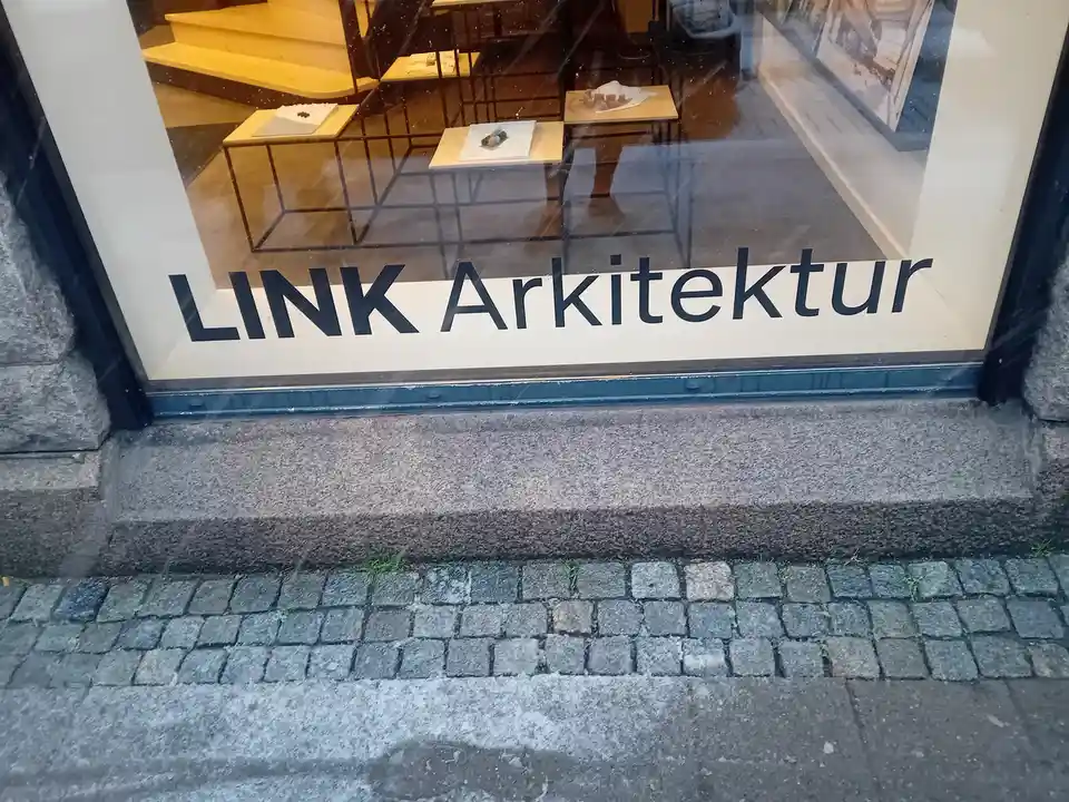 A storefront window displays the text 'LINK Arkitektur' in bold black letters on a white background. Inside, minimalist…