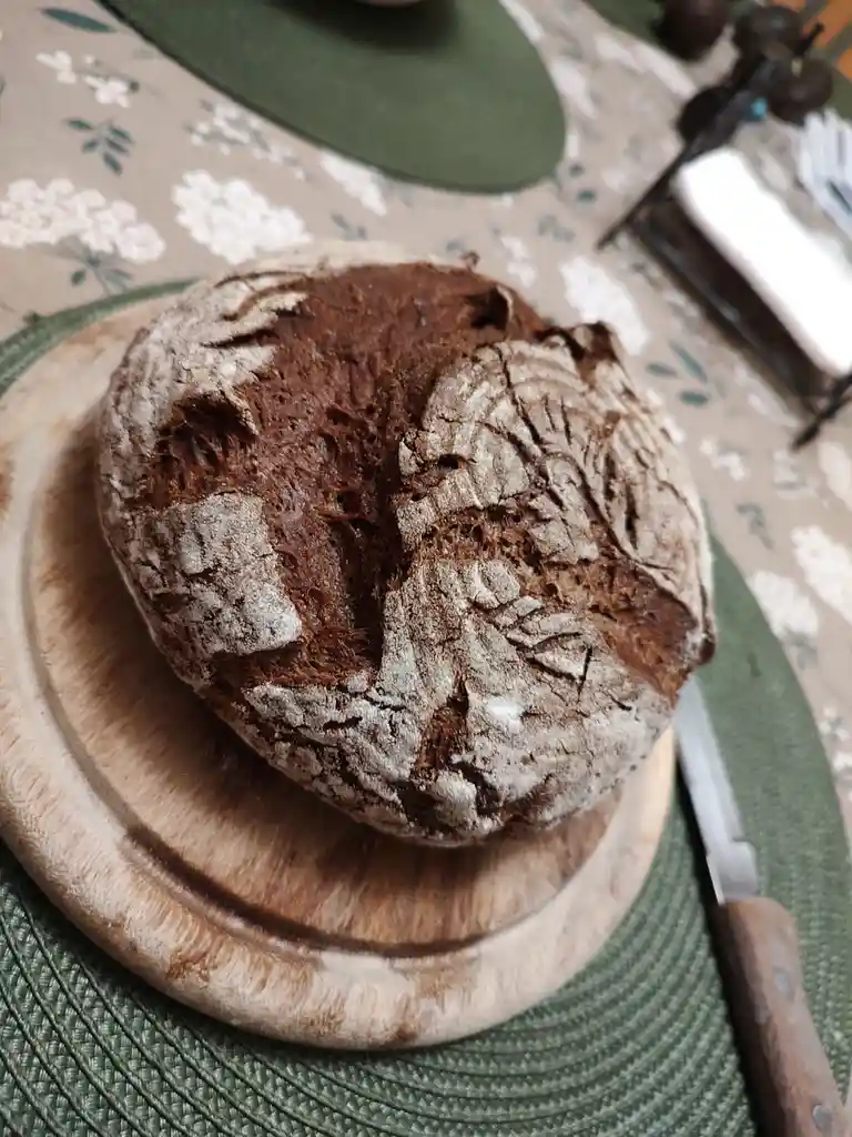 A round, rustic loaf of bread with a cracked, flour-dusted crust sits on a wooden board, its surface resembling a face with…