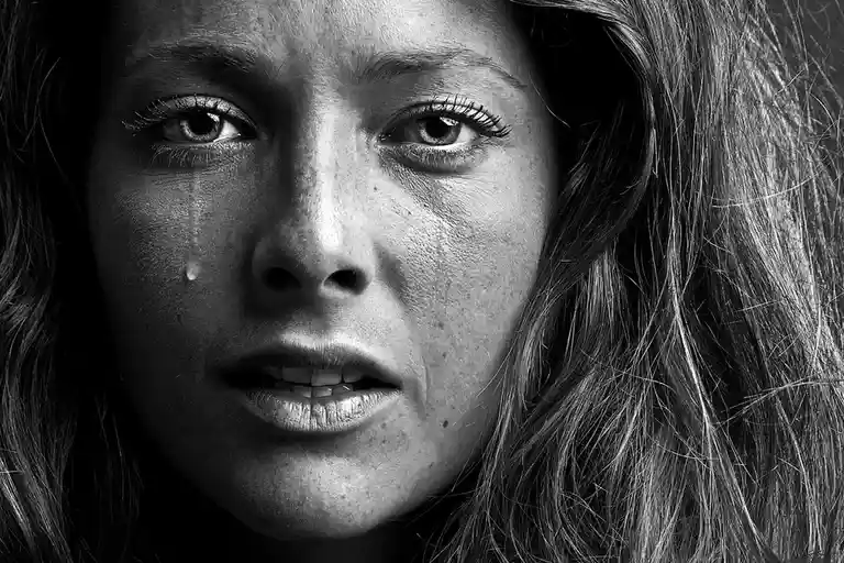 A black-and-white close-up photograph of a woman's face, showing a single tear rolling down her cheek while she stares…