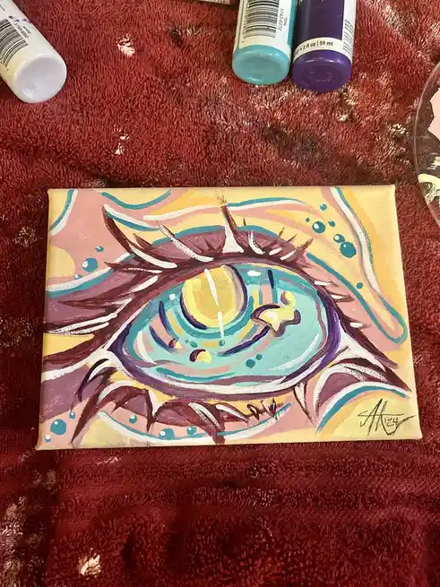A small canvas painting of a stylized, colorful eye with purple eyelashes and teal irises rests on a red towel, surrounded…