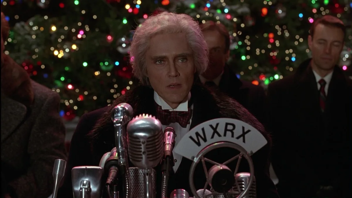 A man with white hair and a bow tie stands at a vintage radio microphone setup with a 'WXRX' sign, looking shocked or…